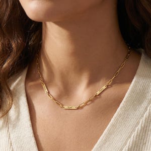 Può includere: Una collana a catena color oro con targhette rettangolari. Le targhette sono incise con i nomi "Karl", "Mary" e "Olivia". La collana è indossata intorno al collo.