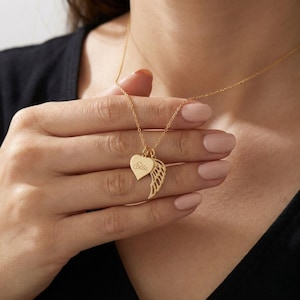 Pode incluir: Um colar dourado com um pingente em forma de coração gravado com o nome "Ella" e um berloque de asa. O colar é exibido em um fundo escuro, destacando o design delicado e o artesanato da joia.