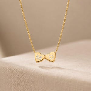 Può includere: Una collana dorata con una catena sottile. Due ciondoli a forma di cuore sono collegati, ciascuno con una lettera all'interno. Il ciondolo di sinistra ha la lettera "M" e quello di destra la lettera "D". La collana è esposta su una superficie di colore neutro.