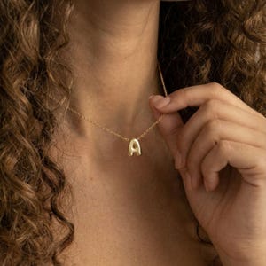 Op de afbeelding: Een goudkleurige ketting met een hanger in de vorm van de letter "A". De hanger heeft een solide, afgerond ontwerp. De ketting is delicaat en heeft een fijne ketting.