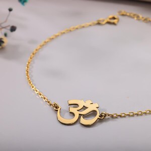 May include: A gold-colored anklet featuring the Om symbol. The anklet has a delicate chain design and a clasp closure. The Om symbol is a prominent, stylized design. The jewelry is displayed on a light gray surface.