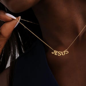 Könnte beinhalten: Eine goldfarbene Halskette mit dem Wort "JESUS" in einzelnen Buchstaben. Die Halskette befindet sich an einer zarten Kette und wird von den Fingern einer Person gehalten. Der Hintergrund ist dunkelblau.