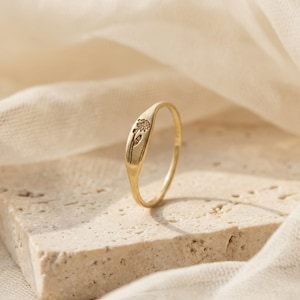 Puede incluir: Un anillo dorado con un diseño floral. El anillo tiene una banda lisa y pulida y una cara rectangular con una flor y un tallo grabados. El anillo se presenta sobre un fondo neutro con una tela suave y fluida.