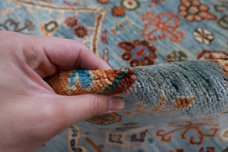 May include: Close-up of a blue rug with intricate floral patterns in shades of orange, brown, and turquoise. The rug is made of a soft, plush material and has a hand-woven texture.