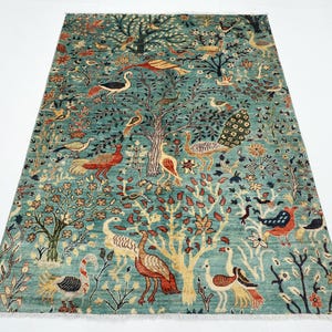 May include: A teal-colored rug with a detailed design featuring trees and various birds. The rug has a central tree motif with peacocks, swans, and other birds scattered throughout the design. The rug has a rectangular shape.