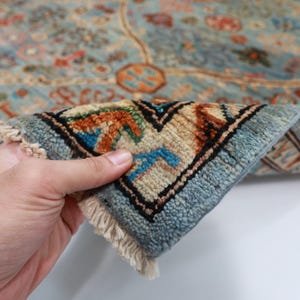 May include: Close-up of a blue and beige rug with intricate patterns. The rug has a fringe border and is folded in half, revealing the underside.