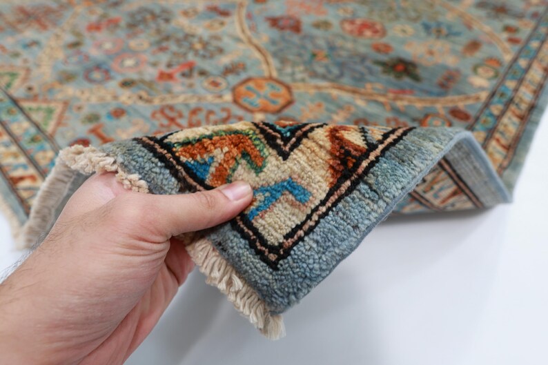 May include: Close-up of a blue and beige rug with intricate patterns. The rug has a fringe border and is folded in half, revealing the underside.