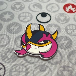 May include: A colorful, cartoonish pin featuring a stylized character with a white face, pink accents, yellow horns, and a navy blue head. The pin is set against a gray background with various symbols.
