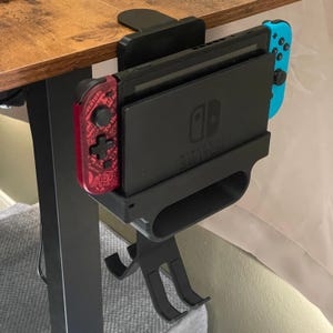 May include: A black desk-mounted Nintendo Switch dock with a red and blue controller attached. The dock is holding a black Nintendo Switch console. The red controller has the text "Adventure" on it.