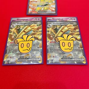Peut inclure: Trois cartes à collectionner Pokémon avec Gimmighoul et Gholdengo EX. Les cartes ont une palette de couleurs or et violet avec un autocollant jaune d'un personnage de dessin animé. Les cartes sont sur une surface rouge.