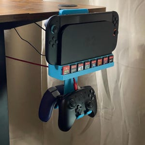 May include: A blue and black gaming console holder attached to a wooden desk. The holder has slots for game cartridges and holds a black console and two controllers. A red charging cable is plugged into the console.