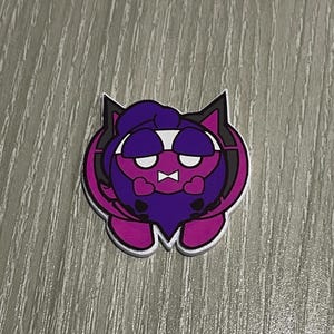 Puede incluir: Un pin de estilo caricatura con una criatura morada con boca en forma de corazón y expresión de mal humor. El pin tiene un borde blanco y está sobre una superficie de madera gris. La criatura tiene barba morada y capucha morada oscura.