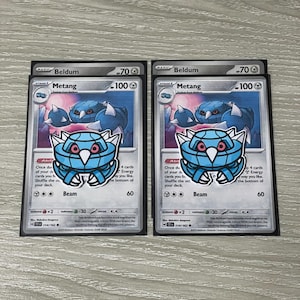 May include: Two identical Metang trading cards, each featuring a blue, crab-like creature illustration with red eyes and white spikes. The cards display the name Metang, 100 HP, and the attack Beam with 60 damage. Each card is inside a protective sleeve.