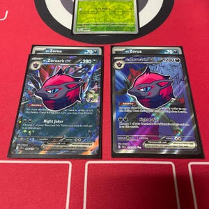 Può includere: Due carte da gioco Pokémon con Zoroark EX. Le carte sono olografiche e presentano uno schema di colori rosso scuro e viola. Il testo sulle carte recita "Night Joker" e "Abilità".
