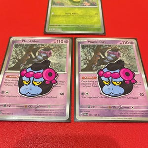 Peut inclure: Trois cartes à collectionner Pokémon. Deux cartes présentent le personnage Munkidori, avec un visage bleu, des cheveux noirs et des accents roses. La troisième carte présente un fond vert avec le texte "Itchy Pollen".