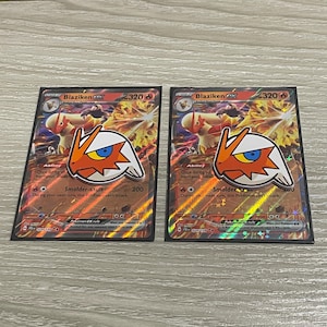 May include: Two Blaziken EX Pokémon trading cards. Each card features a Blaziken character illustration with orange, white, and blue accents. The cards have a holographic background and include text such as "Smolder-sault" and "320".