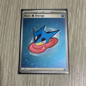 May include: A trading card with a silver border, featuring a blue and pink cartoon character. The card reads "Basic Energy" at the top. The background is blue with a holographic effect. The card is part of a collectible card game.