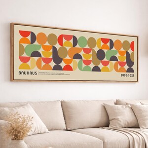 May include: A framed Bauhaus art print featuring a geometric design of colorful shapes in red, orange, green, and brown. The print includes the word "BAUHAUS" and the dates "1919-1933". The artwork is displayed above a beige sofa.