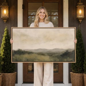 May include: A rectangular landscape painting in a thin wooden frame, featuring a soft, muted depiction of rolling hills and grassy fields under a hazy, pale sky with earthy tones of olive green, beige, and cream throughout the composition.