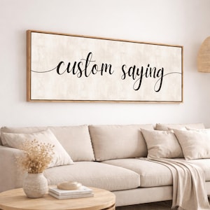 May include: A rectangular wooden framed canvas with the words "custom saying" in black script. The background is a light beige color. The artwork is hanging above a beige sofa.