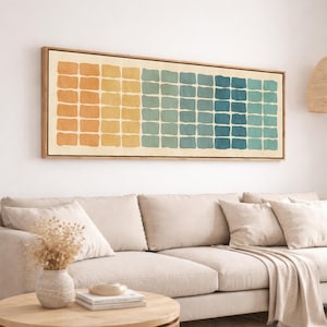 May include: Rectangular wall art with a wooden frame. The artwork features a grid of colorful, square shapes that transition from orange to yellow, then to teal and blue. The artwork is displayed above a beige sofa.