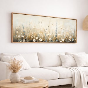 May include: A rectangular framed canvas art piece depicting a field of wildflowers. The painting features white and blue flowers with brown and beige tones. The wooden frame surrounds the artwork, which is mounted on a white wall above a white sofa.