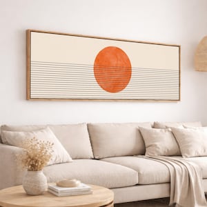 May include: A rectangular wall art piece with a wooden frame. The artwork features a large orange circle above a series of horizontal black lines, set against a cream-colored background. The piece is mounted above a beige sofa.