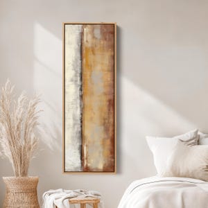 May include: Abstract art piece in a rectangular wooden frame. The artwork features vertical sections of white, gold, and brown hues. The painting has a textured appearance, with visible brushstrokes. The artwork is displayed on a neutral-colored wall.
