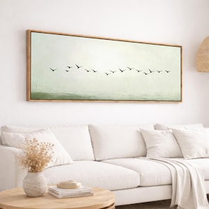 May include: A rectangular framed artwork featuring a flock of black birds in flight against a soft, muted green and white background. The artwork is framed in a light brown wooden frame, adding a touch of warmth to the minimalist design. The artwork is displayed above a white sofa.