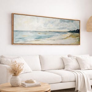May include: A rectangular framed painting of a beach scene. The artwork shows a light blue sea, a sandy shore, and a cloudy sky. The frame is made of light-coloured wood. The painting hangs above a white sofa in a living room.