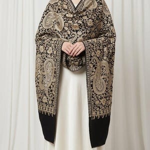 May include: A black shawl featuring intricate gold-toned floral and paisley embroidery throughout the fabric. The rectangular wrap has a dense pattern of swirling botanical motifs and a decorative border along the edges, draped over a plain white dress.