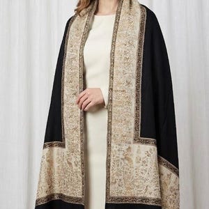 May include: A black shawl with a beige and gold patterned border and accents. The shawl is draped over a cream-colored dress. The beige border features an intricate floral design. The shawl is a luxurious accessory.