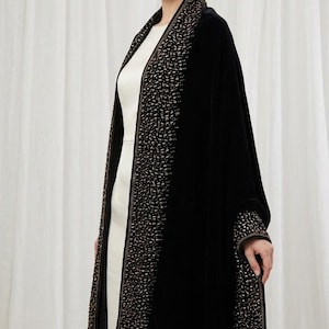 Puede incluir: Un chal de terciopelo negro con un borde ancho adornado con intrincados abalorios metálicos dorados y bronce en los bordes delanteros y los puños, sobre un sencillo vestido sin mangas de color crema con cuello redondo y corte recto.