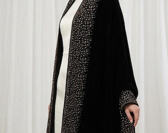 May include: A black velvet wrap featuring a wide border embellished with intricate gold and bronze metallic beadwork along the front edges and cuffs, worn over a simple cream-coloured sleeveless dress with a round neckline and a straight silhouette.