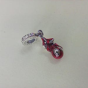 May include: A silver charm with a red, rounded design and a small, oval-shaped detail. The charm has a loop with the word "ERMAN" and "S 925" engraved on it. The charm is likely a jewelry item, possibly a pendant or bead.