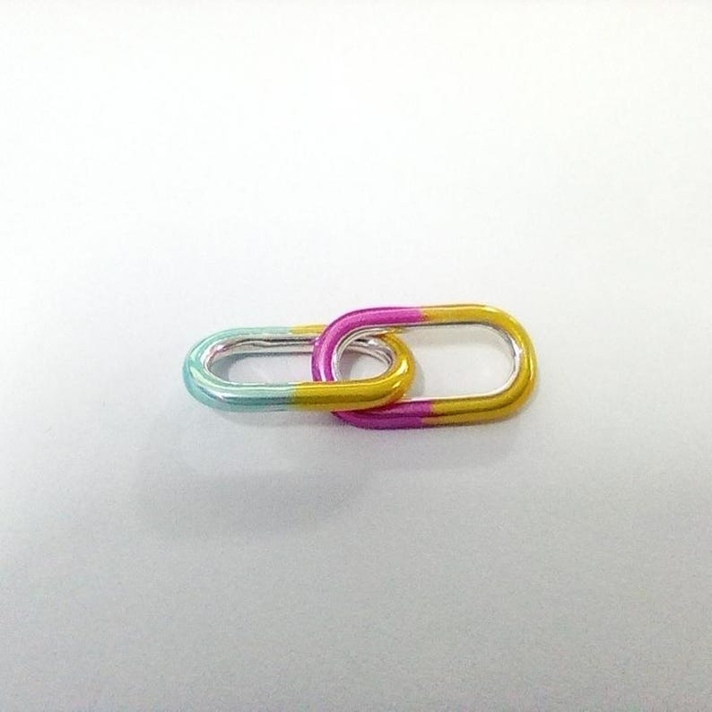 May include: Two linked oval-shaped metal links. One link is teal and silver, transitioning to yellow. The other link is yellow and pink, transitioning to pink. The links are connected, creating a chain-like appearance.