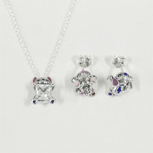 May include: A silver necklace and matching earrings set. The pendant features a square, clear gemstone set within a Stitch-shaped frame with red, blue, and pink accents. The earrings mirror the design, showcasing the same character in a smaller size.