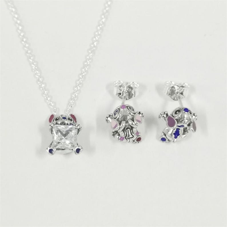 May include: A silver necklace and matching earrings set. The pendant features a square, clear gemstone set within a Stitch-shaped frame with red, blue, and pink accents. The earrings mirror the design, showcasing the same character in a smaller size.