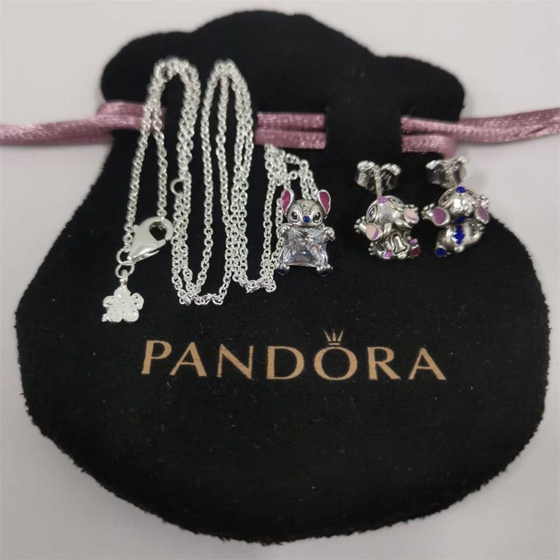 May include: A silver necklace with a flower pendant and a Stitch charm, along with a pair of Stitch stud earrings. The jewelry is displayed on a black pouch with the Pandora logo and a pink ribbon.