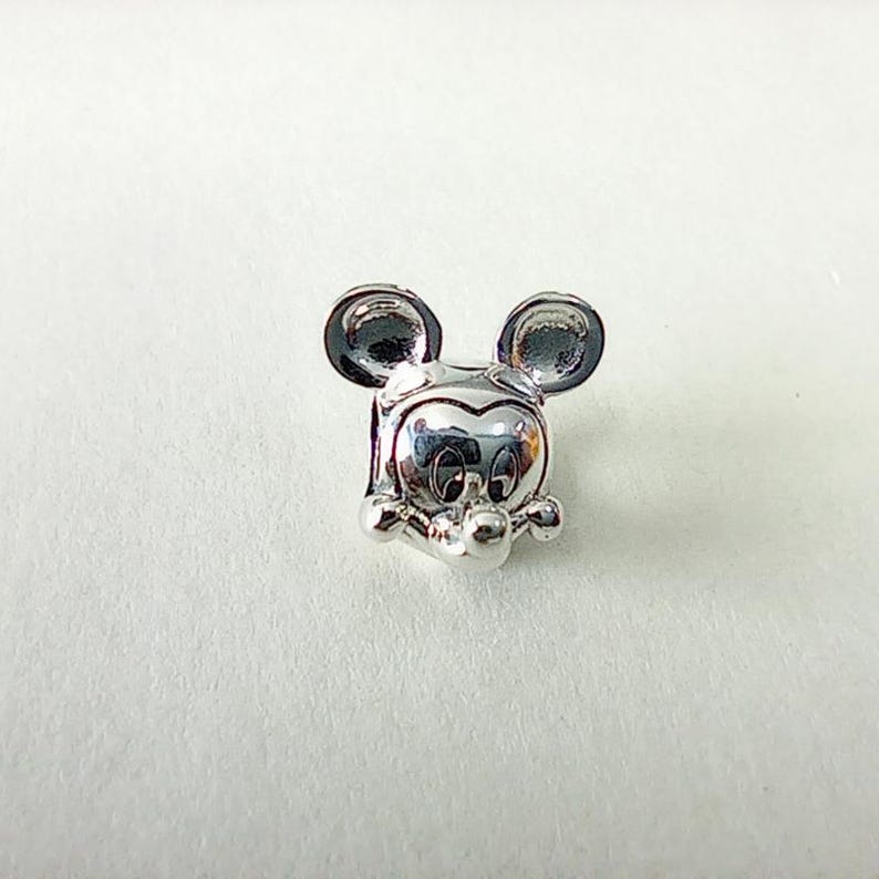 May include: A silver charm featuring the iconic face of a cartoon mouse with large, round ears. The charm has a detailed design with a polished finish, perfect for a bracelet or necklace.