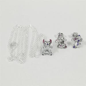 May include: A collection of silver-toned jewelry featuring a necklace with a flower pendant, a pendant of a cartoon character, and two matching character stud earrings. The character has pink ears and blue and purple accents.