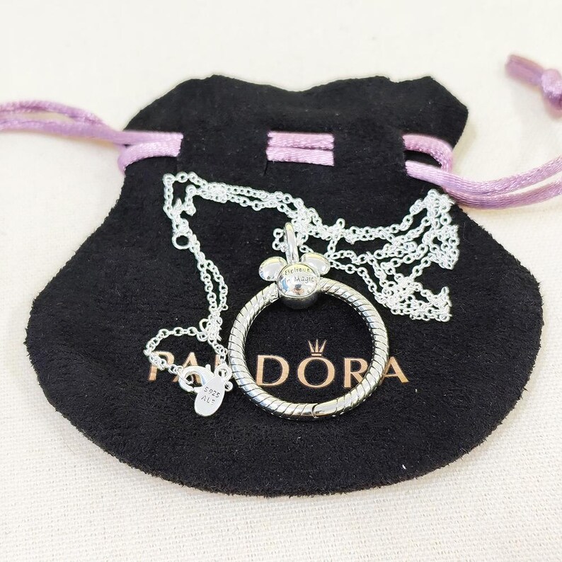 K&ouml;nnte beinhalten: Eine silberne Halskette mit einem kreisf&ouml;rmigen Anh&auml;nger und einem Mickey-Mouse-f&ouml;rmigen Charm. Auf dem Anh&auml;nger steht das Wort "DORA". Die Halskette wird auf einem schwarzen Beutel mit lila Kordelzug pr&auml;sentiert.