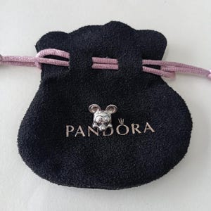 May include: A silver-colored Mickey Mouse charm sits on a black Pandora pouch with a pink drawstring. The pouch has the Pandora logo printed in silver. The charm features Mickey's iconic face and ears.