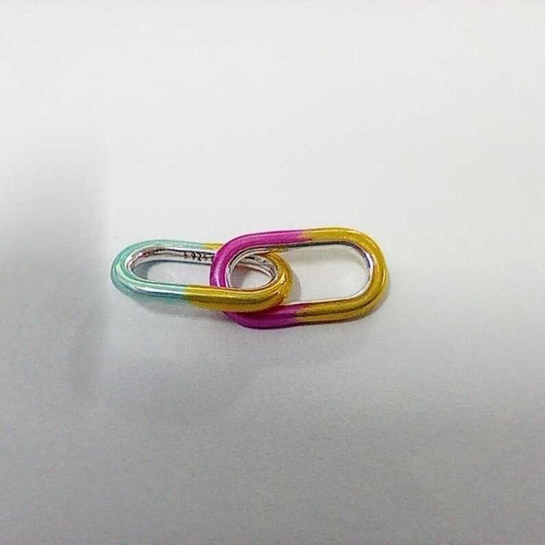 May include: Two linked oval-shaped silver links with a gradient color scheme. One link transitions from teal to yellow, while the other shifts from pink to yellow. The links are connected, creating a unique design.