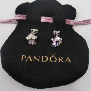 May include: A pair of silver-toned stud earrings featuring a cartoon character design with pink, purple, and blue enamel accents. The earrings are displayed on a black velvet pouch with the "PANDORA" logo.
