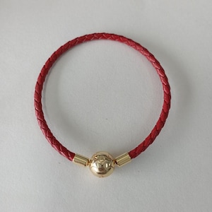 Puede incluir: Una pulsera de cuero trenzado rojo con un dije esférico dorado. La pulsera es circular y parece estar hecha de cuero genuino. El dije dorado tiene un patrón decorativo y se sujeta con cierres metálicos dorados.