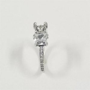 May include: A silver ring featuring a stylized animal figure with large eyes and ears, holding a square, clear gemstone. The ring band is adorned with small, sparkling stones, creating a delicate and eye-catching design.