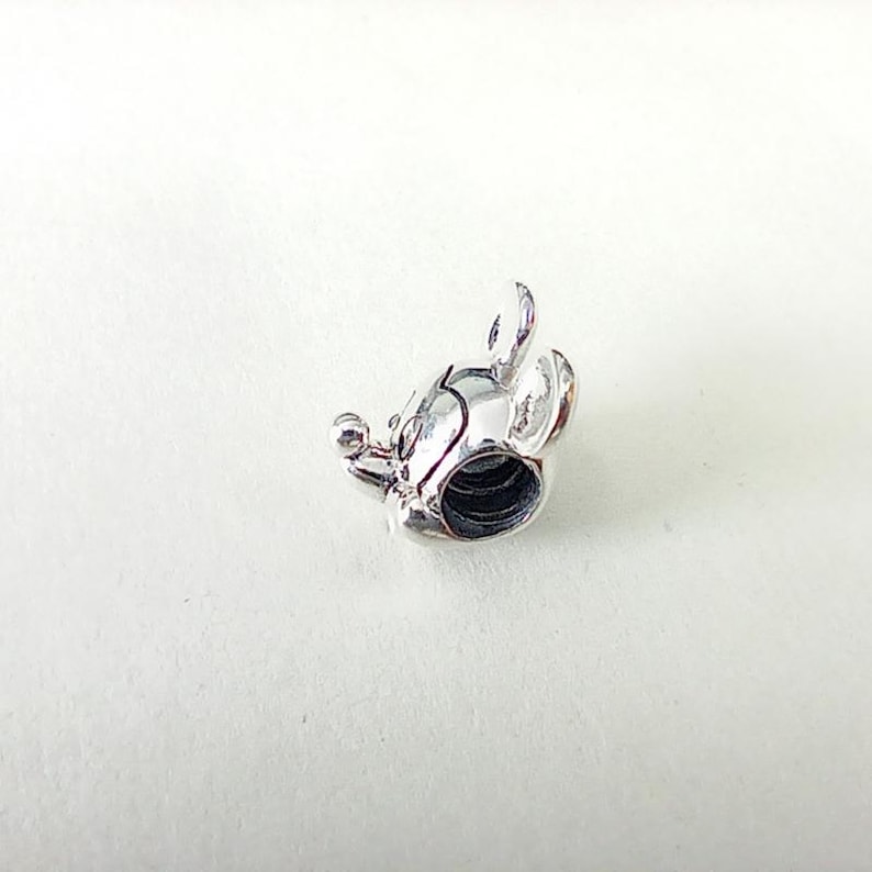 May include: A silver-colored charm in the shape of a mouse's head, featuring rounded ears, a small nose, and a circular opening. The charm is highly detailed and appears to be made of metal. The design is reminiscent of a popular cartoon character.
