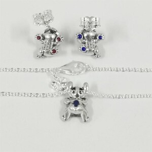 May include: Silver-toned jewelry set featuring a charm necklace and matching stud earrings. The charms and earrings are shaped like a cartoon character with red and blue accents. The necklace has a delicate chain and a lobster clasp.