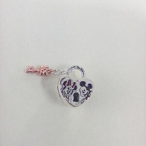 May include: A silver heart-shaped padlock charm with a rose gold key. The padlock features engraved images of Minnie and Mickey Mouse. The key is attached to the padlock with a small chain. The charm is on a white background.
